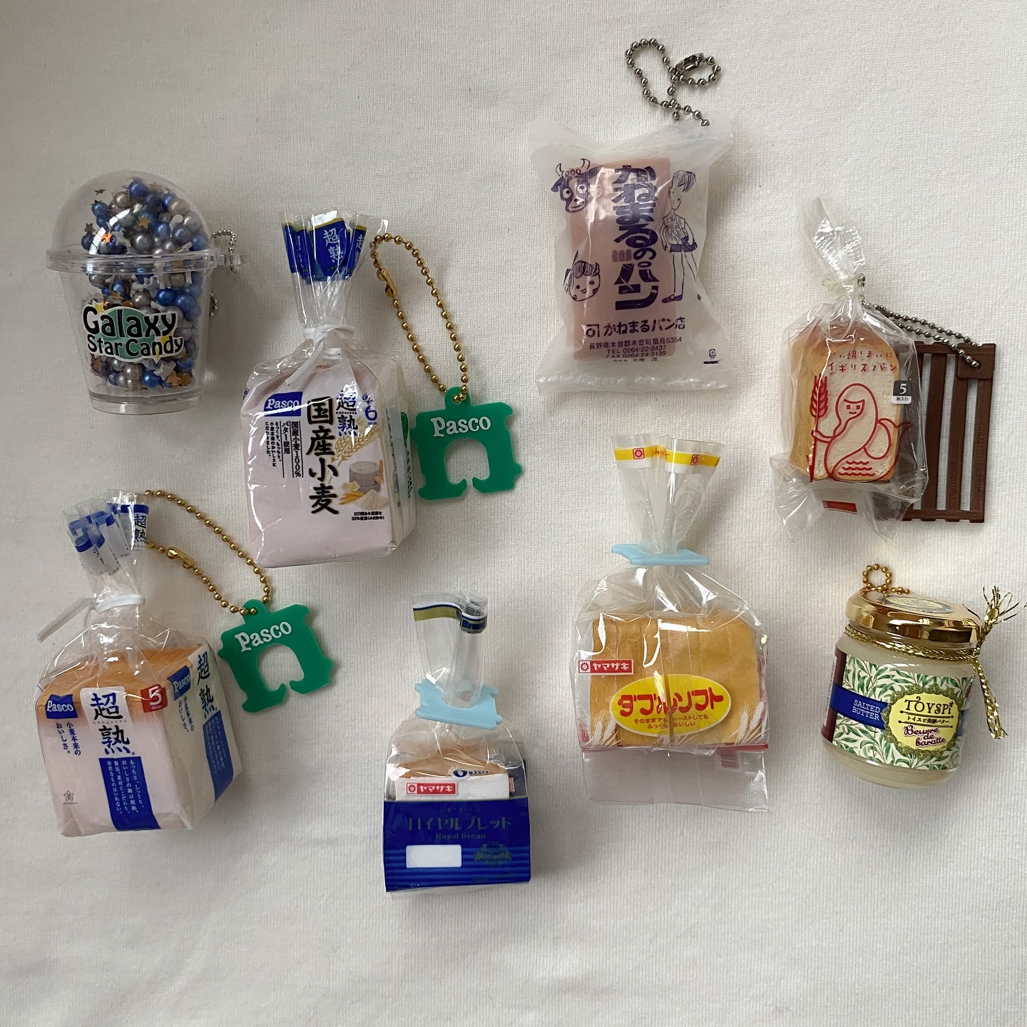 the image displays a collection of miniature food keychains and toys arranged on a white surface. prominently featured are several bread keychains, each replicating different brands of japanese bread such as pasco, yamazaki, and kakumaru. each bread keychain is packaged in clear plastic, sealed with a twist tie and connected to a metal ball chain. two of the pasco bread keychains have a green pasco tag. other items include a "galaxy star candy" keychain in a clear plastic cup and a toy jar labeled "toyspy". the arrangement suggests a collector's display or a selection of novelty items.