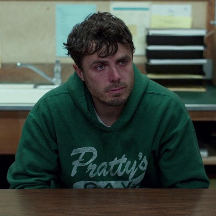 a man sits alone at a table looking down, wearing a green hoodie with the words "pratty's" on it. he looks to be in his 30s and has a serious expression. his face is partially obscured by his hair, and his facial expression suggests that he might be sad or worried. he is the only person in the image.