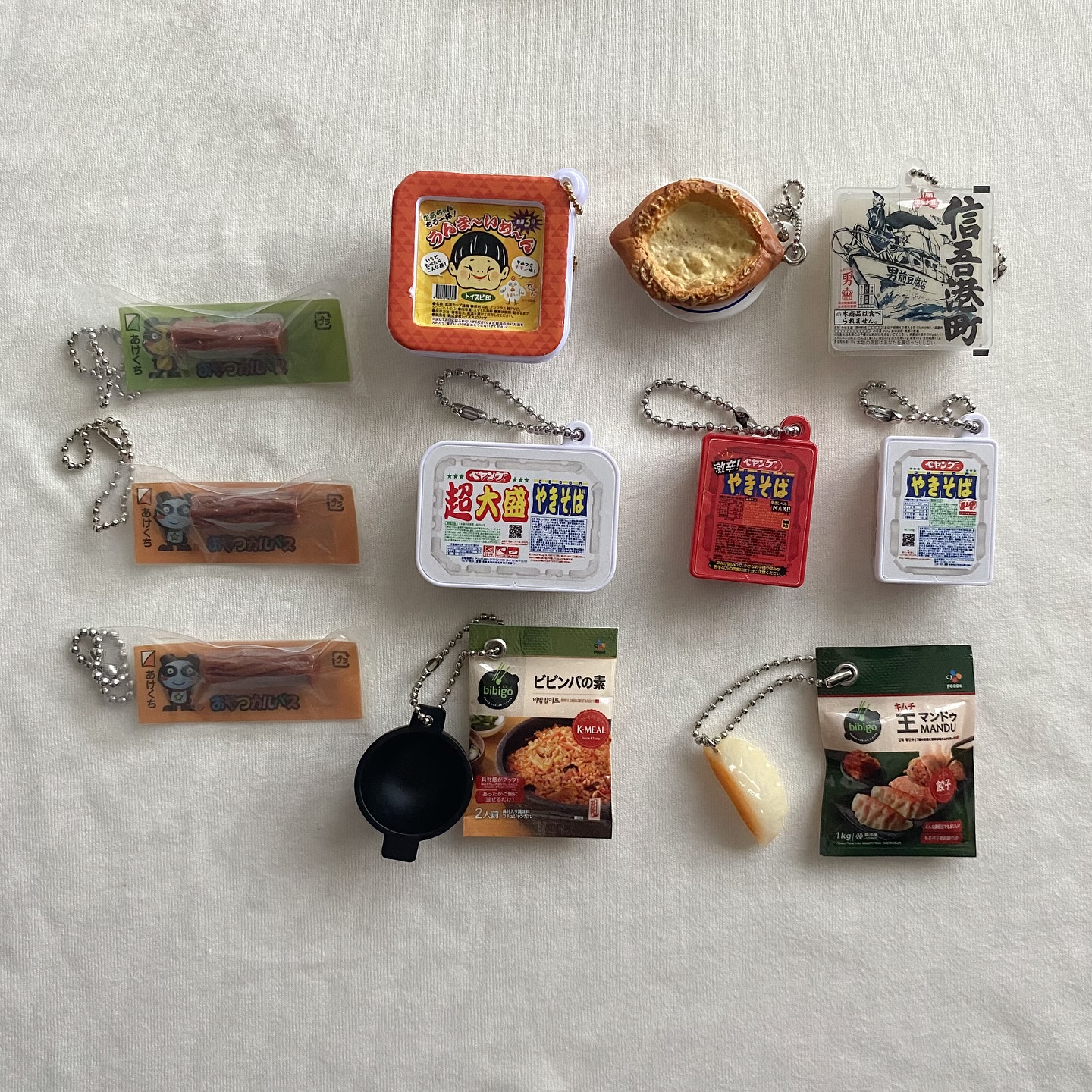 the image shows a collection of foodthemed keychains laid out on a white surface. there are several representing japanese cuisine, including yakisoba in different packaging colors (white, red), tsukutsukutsukun (sausage snack) and an anime character on a square packaging. additionally, there are keychains of korean foods like bibigo bibimbap and wang mandu (dumplings), along with an egg tart and a mini frying pan. each keychain has a silver ball chain attached to it.