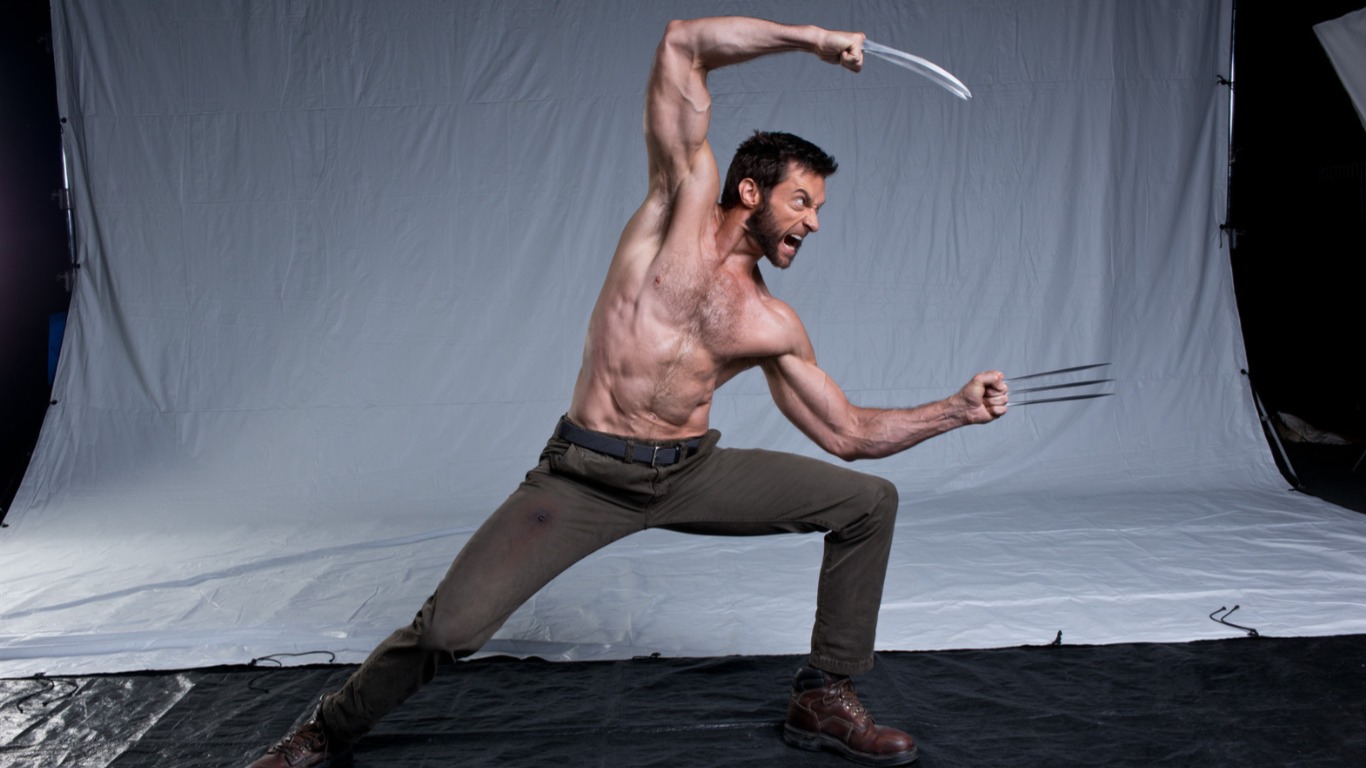 a shirtless hugh jackman as wolverine in an action pose, wielding claws. he's wearing brown pants and boots in a studio setting against a white backdrop. he is scowling aggressively.