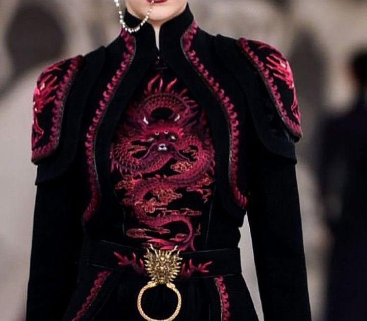 a detailed shot of a woman's upper torso wearing a black dress with intricate red dragon embroidery on the front and shoulders. the dress has a high collar and is adorned with shoulder pads. a belt with an ornate gold buckle featuring a ring detail cinches the waist. the overall aesthetic is high fashion and draws inspiration from asian design.