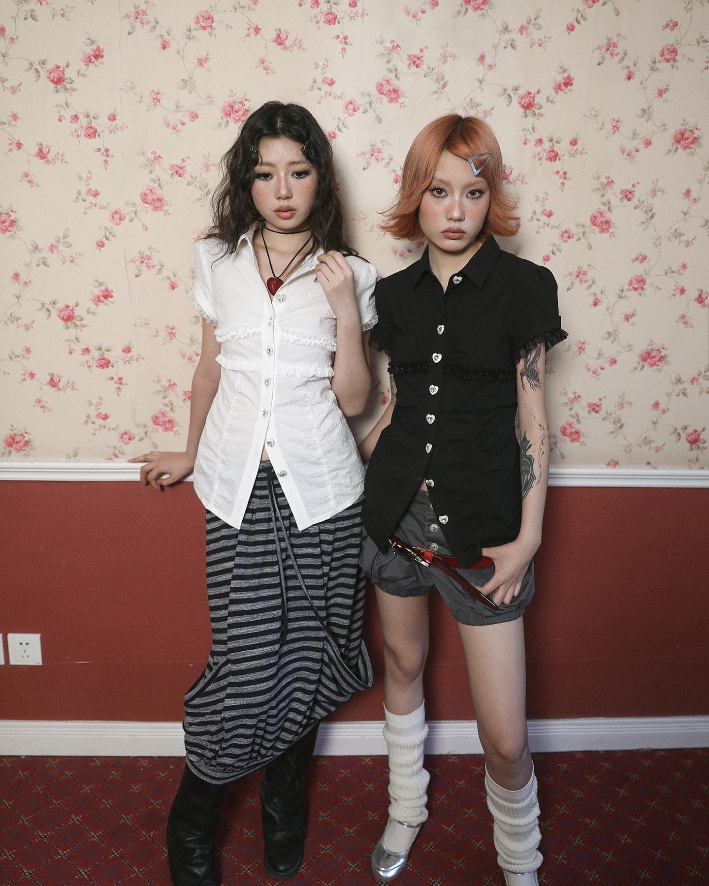 two young women are standing against a floral wallpapered wall. one is wearing a white ruffled shirt and a striped maxi skirt, while the other wears a black buttonup shirt and short shorts. both are wearing white socks and shoes.  the setting appears vintage or retro, with a red wall and ornate baseboard.  they are posing for a photo.