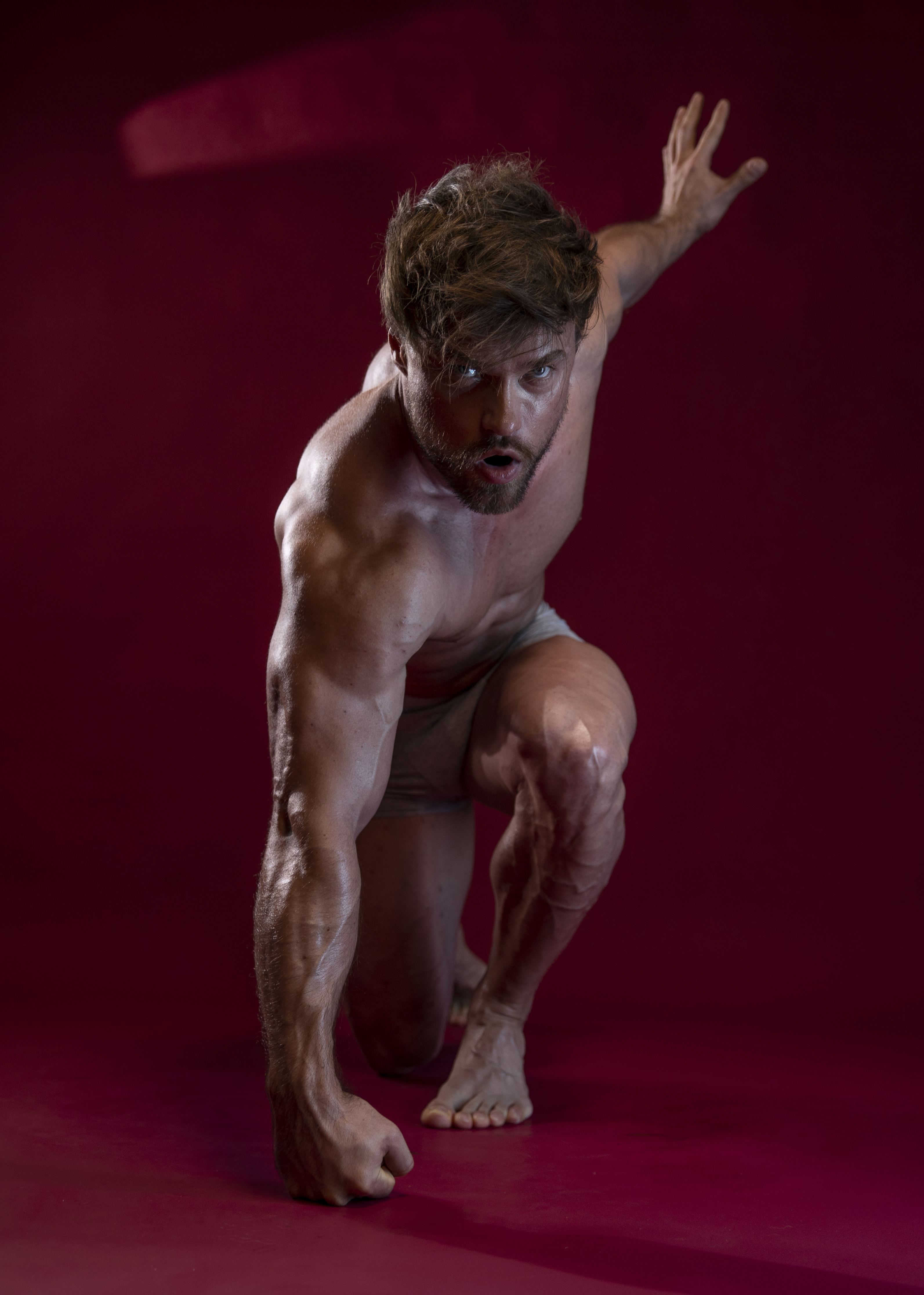 a muscular man is kneeling shirtless against a vibrant red background. he is a bodybuilder, showcasing his toned physique and prominent veins. he poses dynamically with one arm extended and an intense gaze directed towards the viewer. the lighting accentuates his muscularity and athletic build. he has a beard and his hair is tousled.