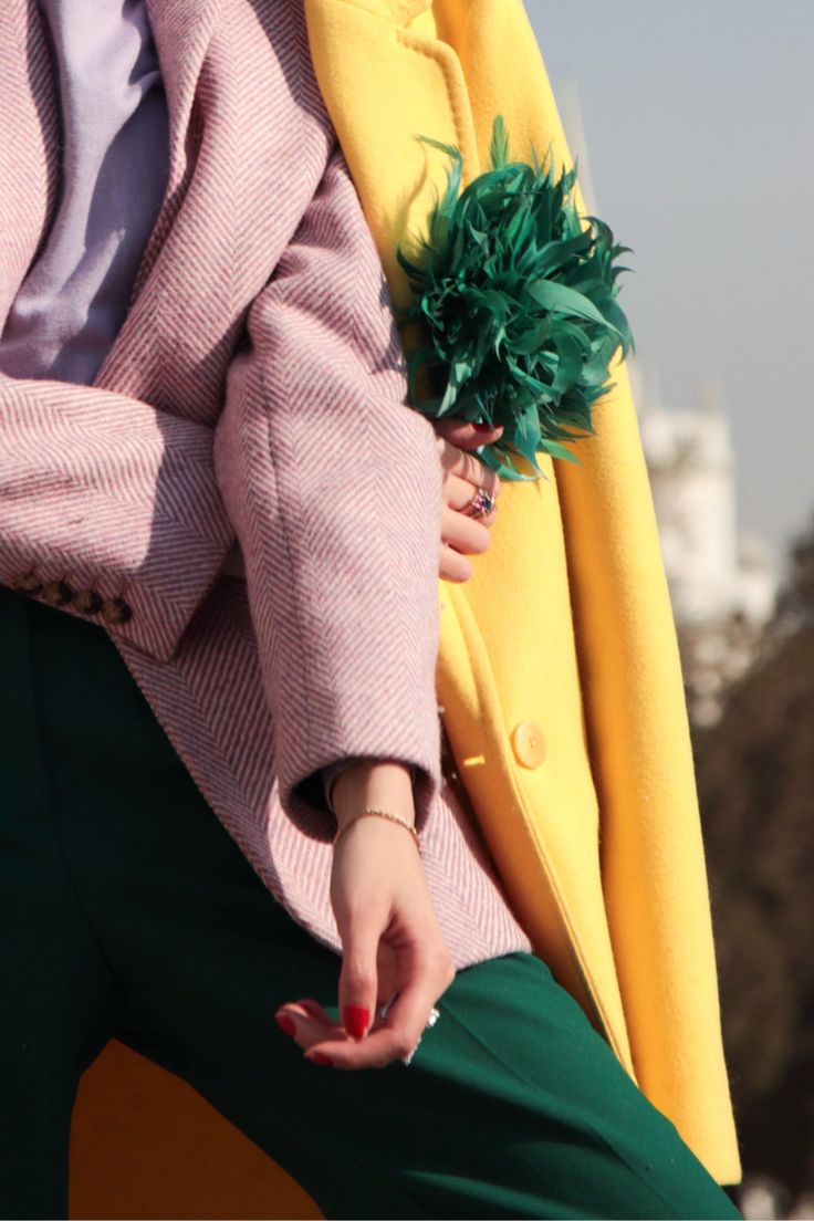 closeup shot of a woman's stylish outfit. she's wearing a pink herringbone blazer over a lavender top and emerald green trousers. a bright yellow coat is draped over her shoulders. a striking green feather brooch is pinned near the collar. the woman's hand is visible, showcasing red nail polish and jewelry. the background is softly blurred, suggesting an outdoor setting in natural light. the overall image conveys a sense of fashion and sophistication.