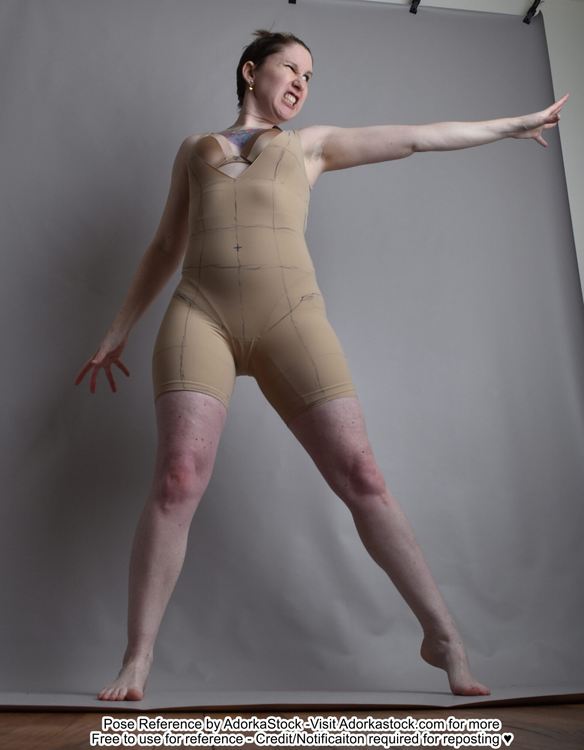 a woman in a nudetoned body suit stands with her legs spread and her left arm outstretched, reaching toward the camera. her right hand is clenched into a fist. she is looking towards the camera with an angry expression.  her  dark, brunette hair is pulled up off of her neck.  the woman is standing in a studio with a gray background. the picture is taken from a low angle, and the woman’s feet are in focus.