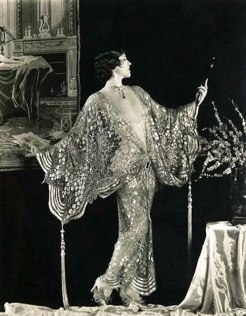 a black and white portrait of a woman in the 1920s. she is wearing a lace robe and jewelry, holding a cigarette holder. she is posing in an elegant interior setting with a decorated fireplace and a table with flowers. her fashion and posture showcase the glamour of the era.