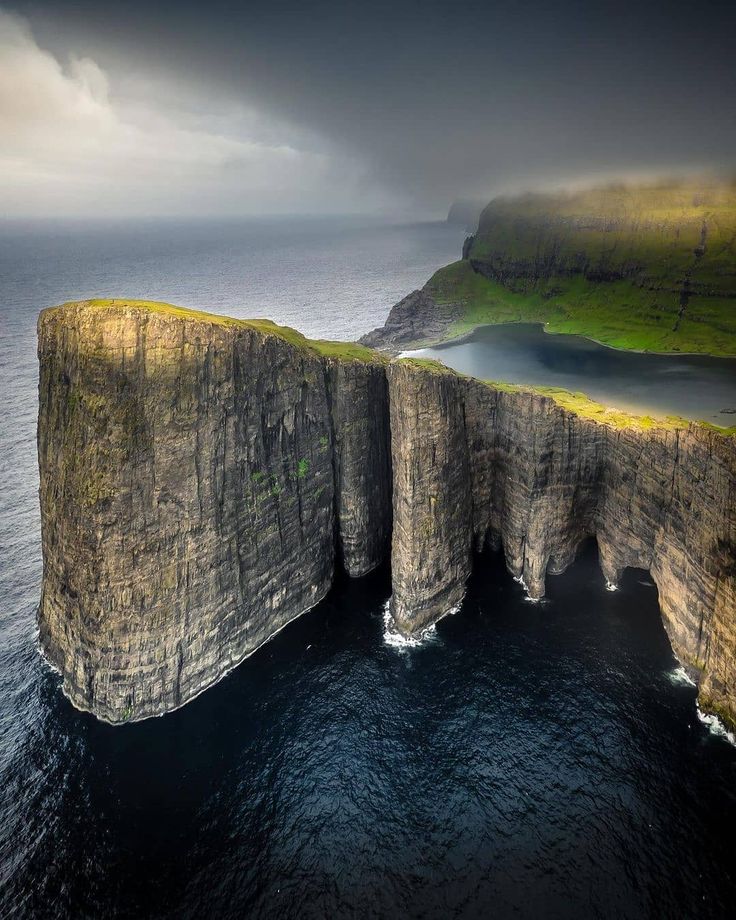 an aerial view of the stunning vestmanna bird cliffs in the faroe islands. the dramatic cliffs rise up from the ocean, with their rugged faces and deep crevices. the sky is overcast with clouds, adding to the mood of the scene.  the sea below is a deep blue color, and the cliffs are covered in green vegetation.  the iconic lake in the cliffs is also visible in the image.  the image is a testament to the raw beauty of the faroe islands.