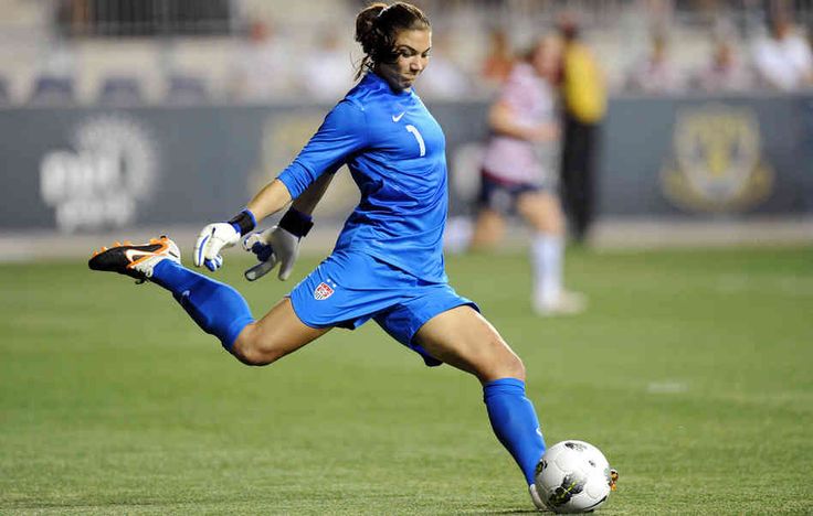 a female soccer goalkeeper in a blue uniform leaps to her right, her left leg extended and right leg bent.  she's contacting the soccer ball with her right foot.  she's wearing gloves and has her hair pulled back. the background is blurred and shows other players and a green soccer field.