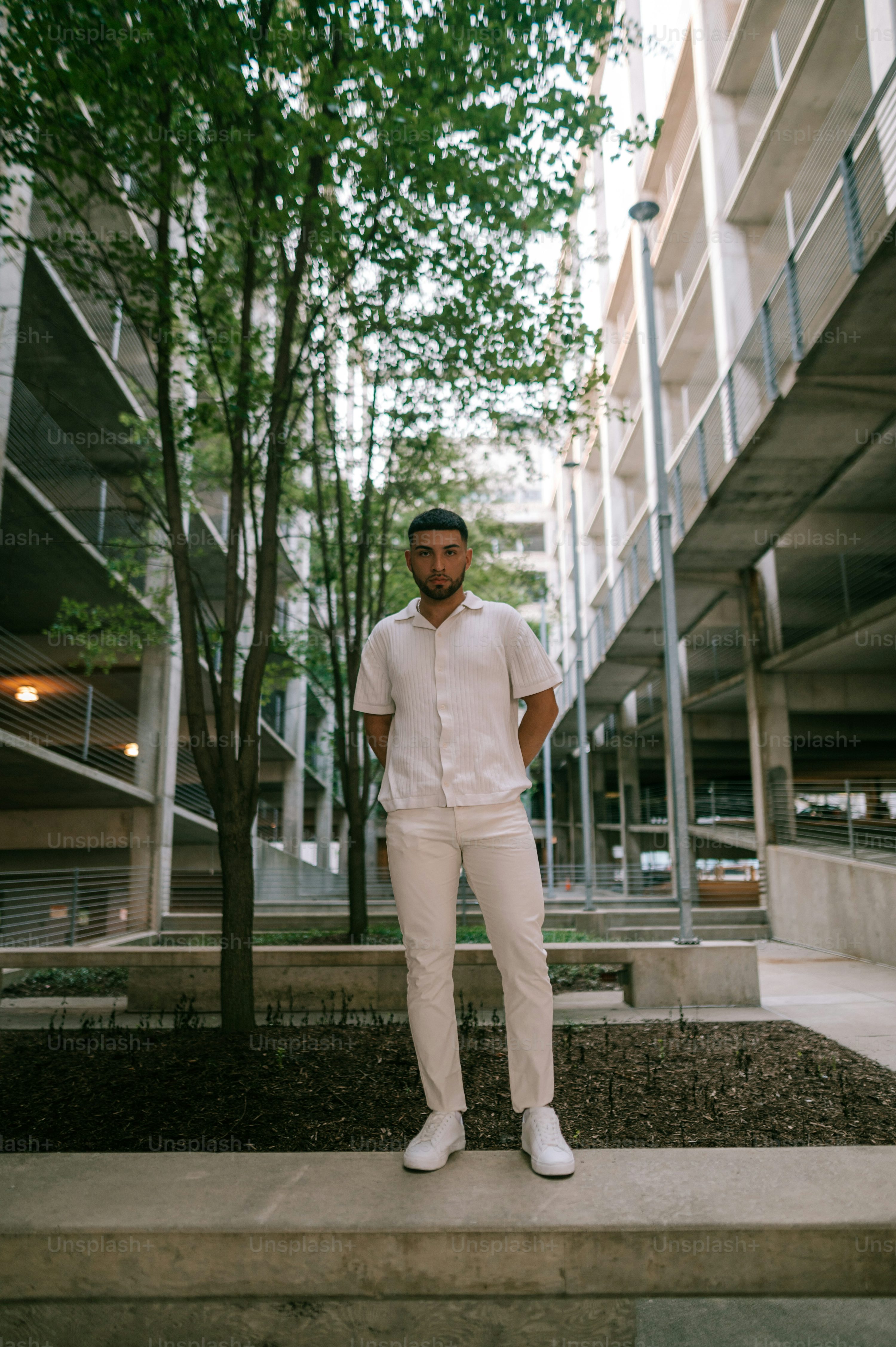 a young man is standing in the middle of a modern, urban setting, specifically what appears to be a parking garage with walkways and green landscaping. he is wearing an allwhite outfit that includes a buttondown shirt, pants, and sneakers.  the style is minimalist and clean. he stands with a serious expression, and his pose is confident. the background features tall concrete buildings and trees. the overall aesthetic is contemporary and somewhat monochromatic due to the prevalence of white and neutral tones.