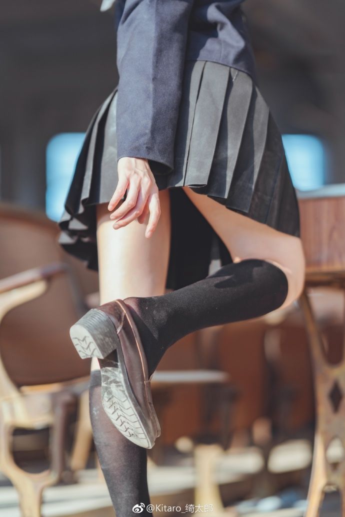 the image shows a woman wearing a school uniform with a pleated skirt, kneehigh socks, and brown shoes. she is sitting on a desk, with one leg extended. a chair is partially visible in the background.