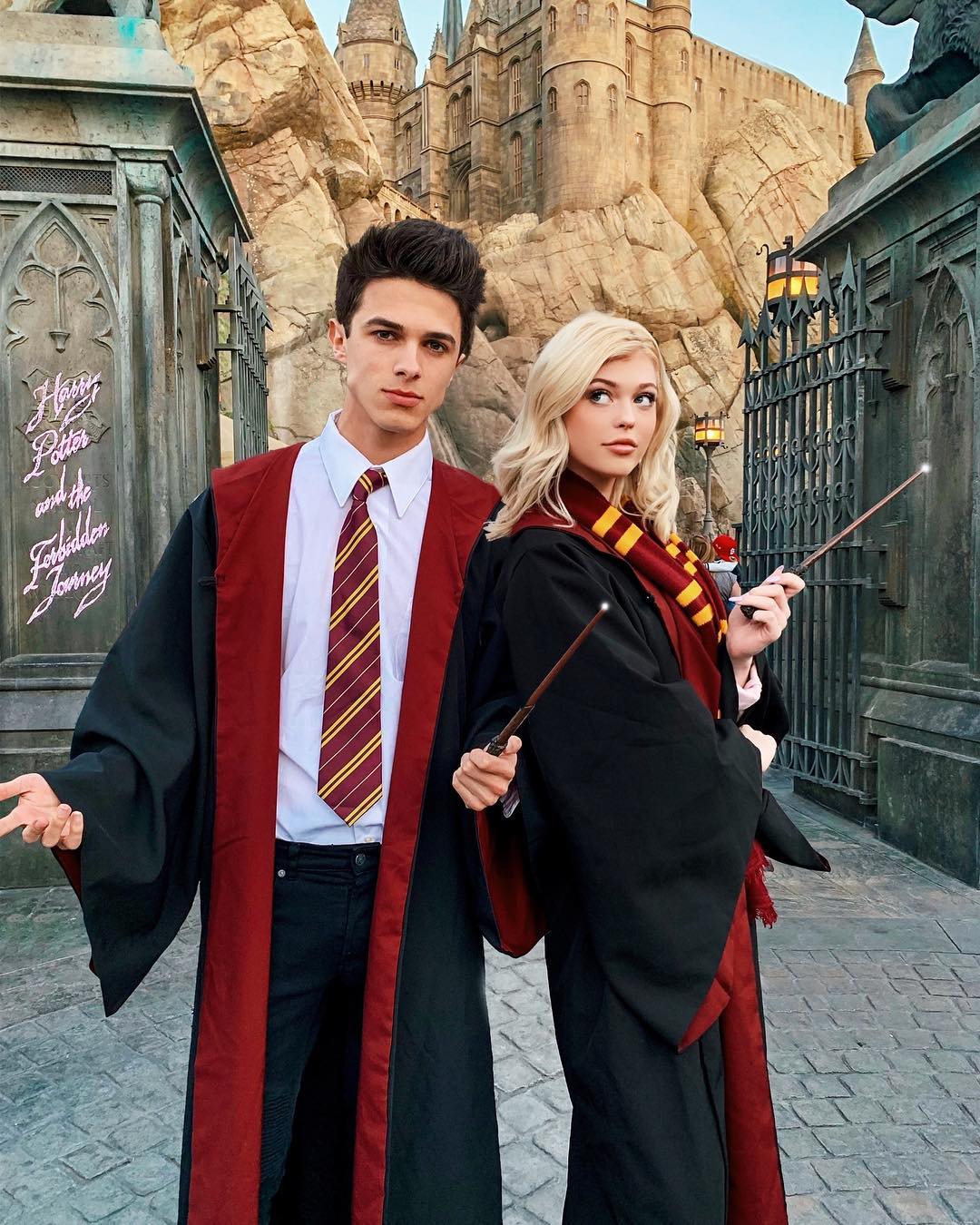 a man and a woman dressed as harry potter characters are posing with wands in front of a hogwarts castle backdrop. they are standing by a gate at a theme park. the man wears a white shirt, tie, black jeans, and a gryffindor robe, while the woman wears a black robe with a gryffindor scarf. they are holding wands and appear to be casting spells. the background includes a castle, rocky terrain, and a sign for "harry potter and the forbidden journey".
