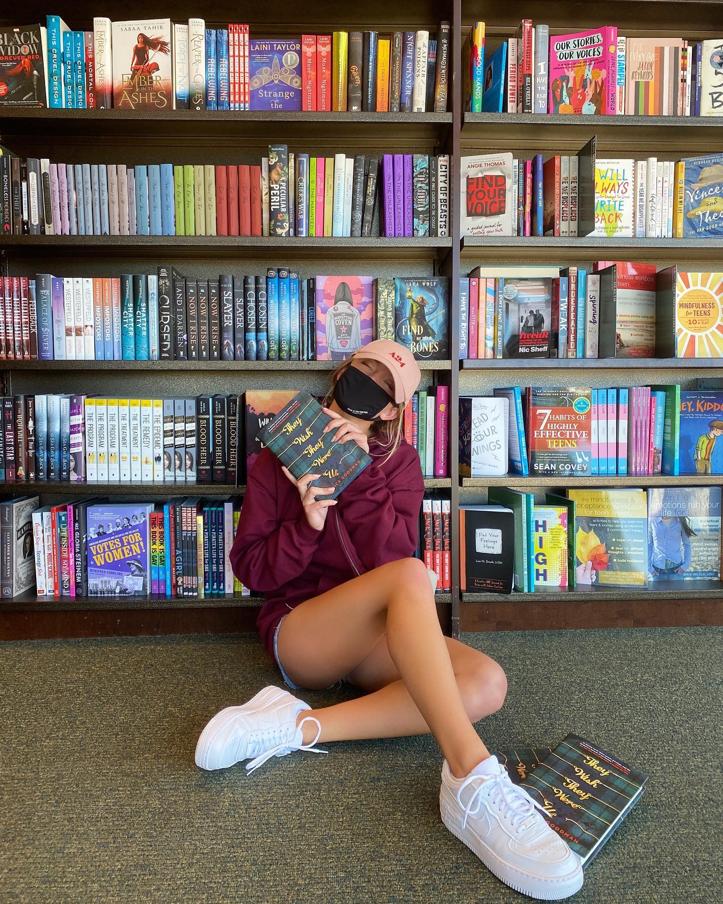 the image shows a young woman sitting on the floor of a bookstore surrounded by numerous bookshelves filled with various books. she is wearing casual attire including white nike air force 1 sneakers and a maroon sweatshirt. she is holding a copy of the book "they both die at the end," and another copy is visible on the floor near her feet. the setting is warm and inviting, typical of a wellstocked bookstore. the overall mood is relaxed and literary.