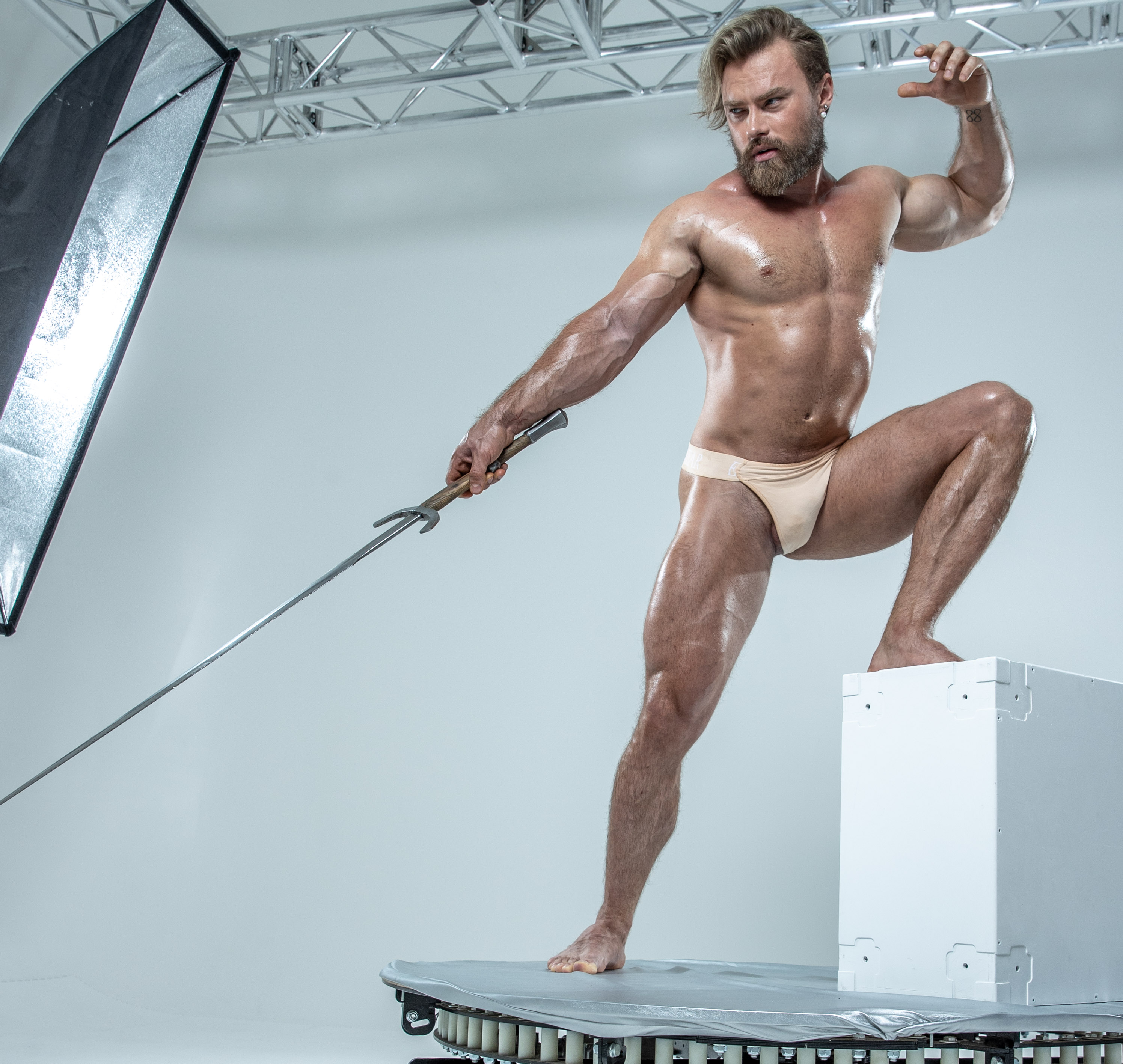 a muscular, bearded man in white underwear strikes a dynamic pose on a white platform in a studio. he holds a sword confidently, his body glistening with sweat. a large lighting reflector is positioned to the left, highlighting his physique. the background is a clean, bright white, emphasizing the man and his powerful stance.