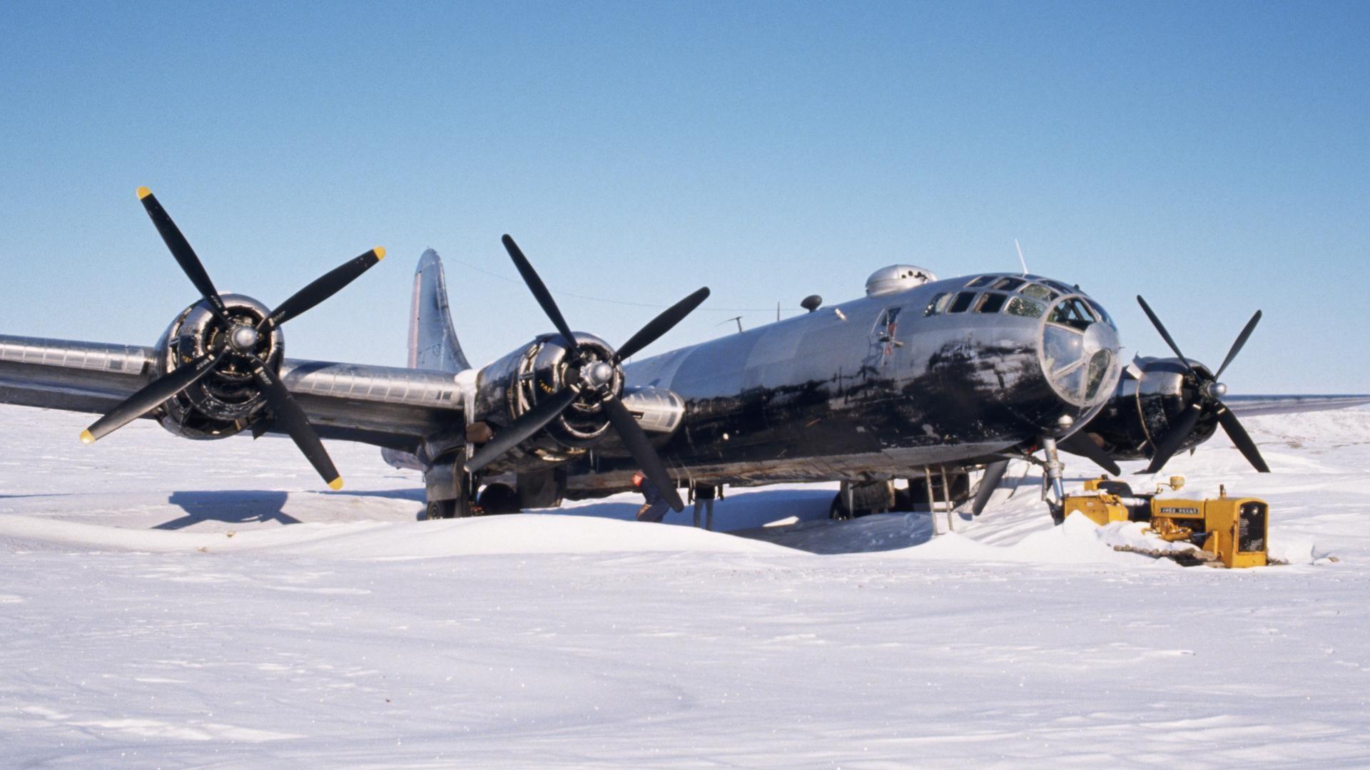 a b29 superfortress bomber is parked on a snowcovered runway. the bomber is painted in a silver and black scheme, and the propellers are black. the aircraft is in the process of being towed by a yellow tractor.