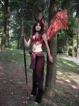 a woman in a vibrant cosplay costume, meticulously designed to portray a fantasy demon or devil character.  she's wearing a red and maroon outfit, complete with horns, large red wings, and dark boots.  the cosplay incorporates details like a staff or spear, adding to the fantasy aesthetic.  she is standing in a forest setting, posing for a photograph. the image suggests a character from a video game or fantasy novel.