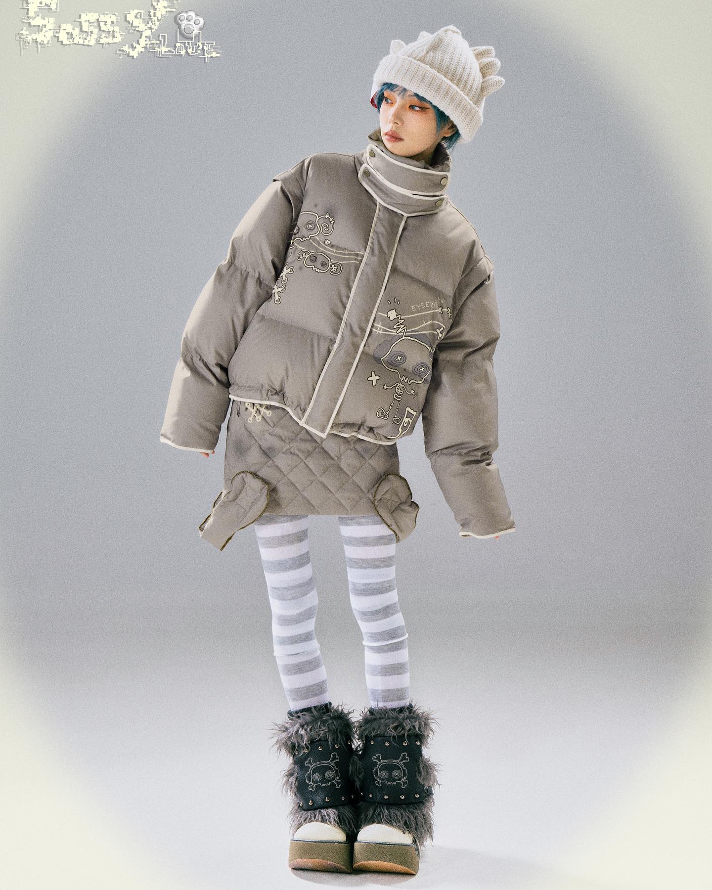 the image shows a fullbody shot of a model wearing a stylish winter outfit. the focal point is a light grey oversized puffer jacket adorned with cartoon embroidery.  the jacket is paired with a white knit beanie, striped tights, and dark grey furry boots featuring a skull motif. the model is posing against a light grey background, showcasing the outfit's details. the overall aesthetic is a blend of kawaii and streetwear influences, possibly leaning towards lolita fashion, with its playful and whimsical details.