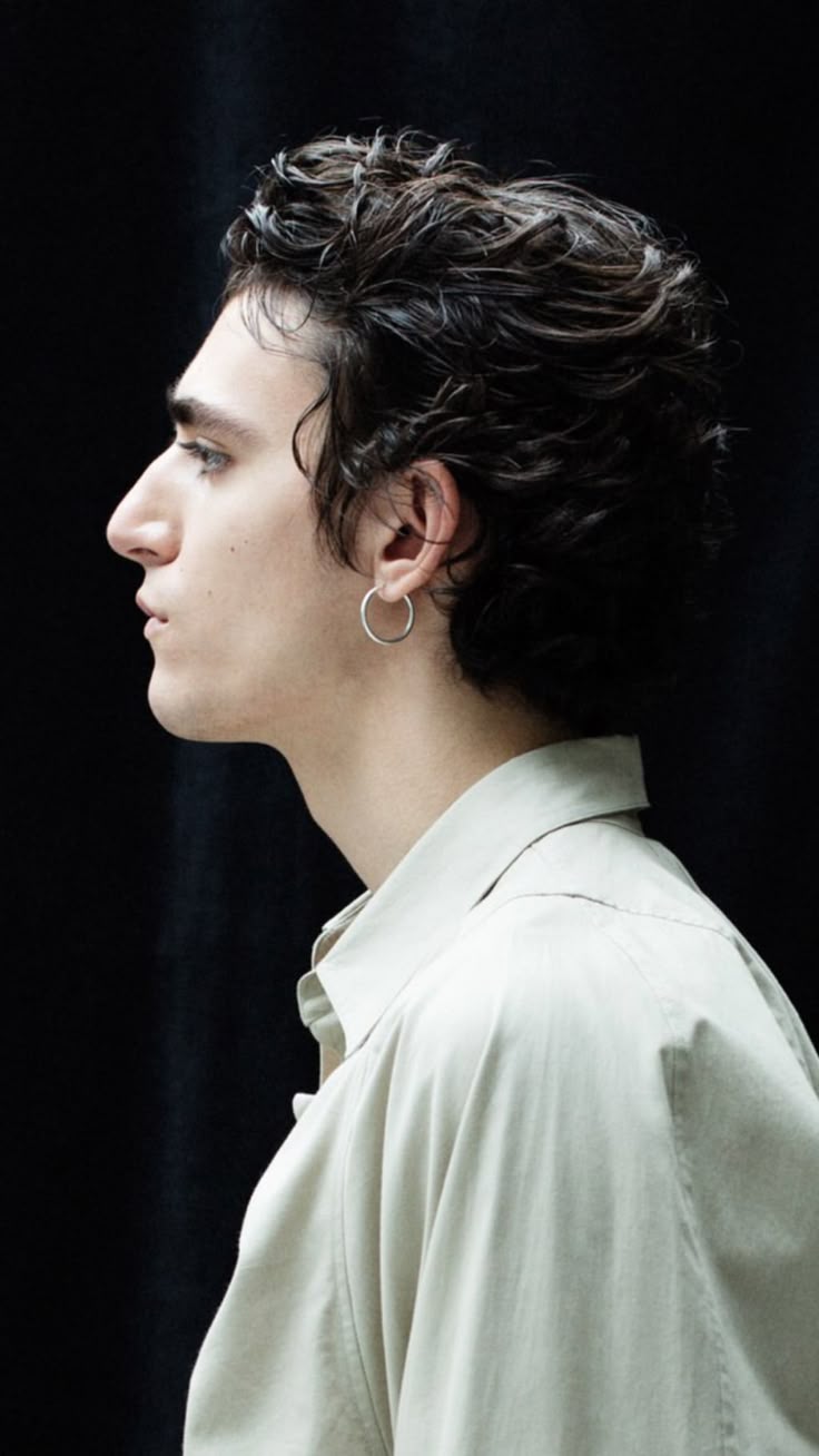 a profile portrait of a man with dark, wavy hair and a silver hoop earring. he is wearing a lightcolored shirt and is set against a black background.