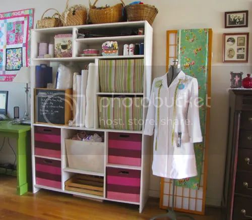 a brightly lit sewing room with a white shelving unit full of baskets, storage bins, fabric, and other sewing supplies.  a sewing dress form with a white jacket, a folding vintage screen, a desk and sewing supplies.  there is a quilt on the wall.