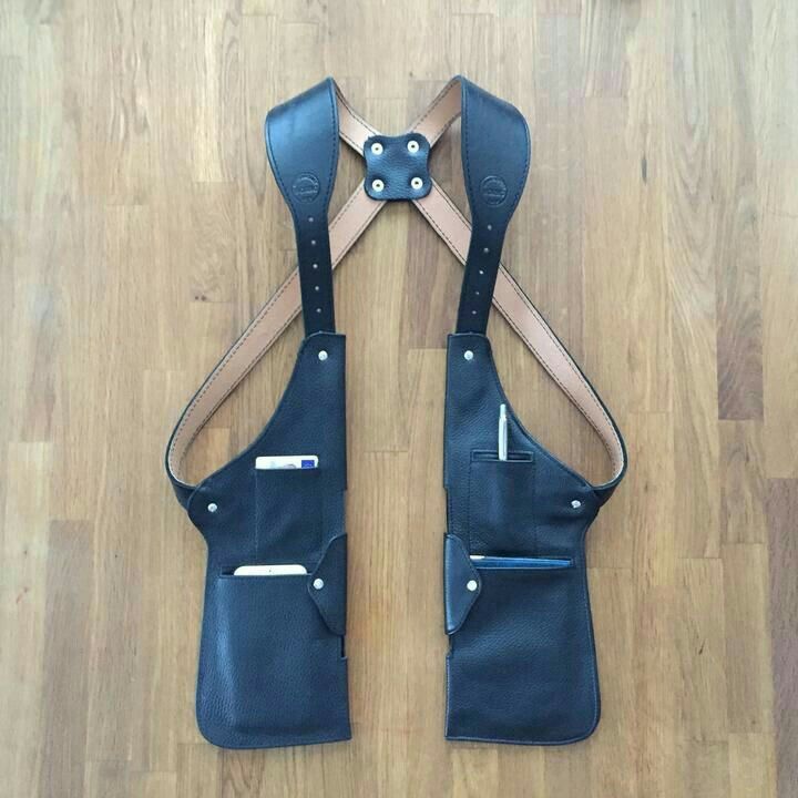 a black leather shoulder holster with tan straps is laid out on a wooden floor. the holster has pockets on each side and is designed to be worn over the shoulders. the straps are tan and are connected at the back with a leather xshaped piece.