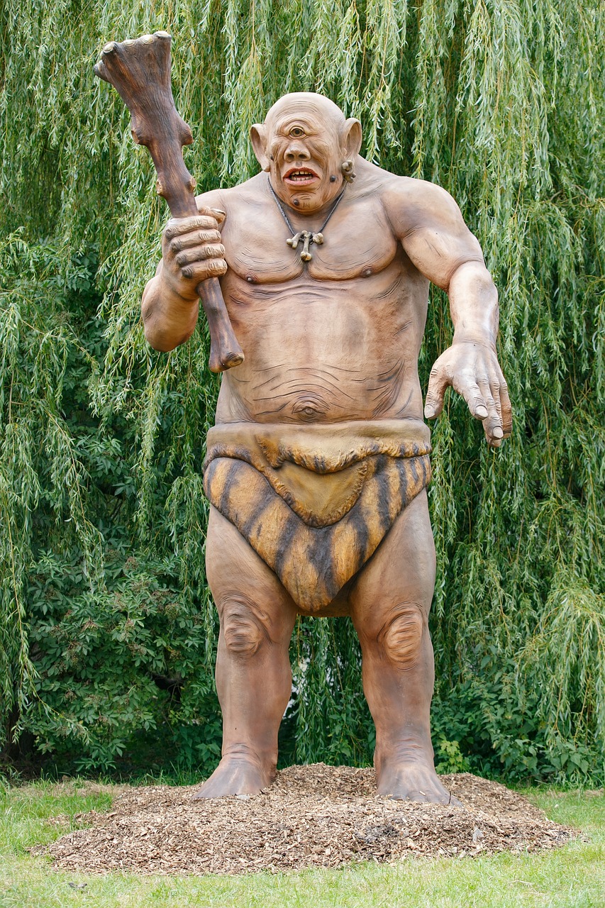 a full shot of a cyclops statue standing in front of a tree. the statue depicts a muscular, oneeyed giant holding a club. it is standing on a pile of wood chips. the tree behind the statue has dense foliage. the statue is an artwork of a mythological creature.