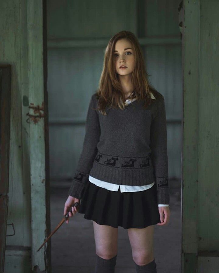 a young woman is dressed in a harry potterinspired schoolgirl outfit, including a grey vneck sweater with an embroidered design on the bottom, a white buttondown shirt, a black pleated skirt, and grey kneehigh socks. she is standing in the doorway of a dimly lit, seemingly abandoned room and is holding a wand in her right hand. she has long, straight reddishbrown hair and is looking directly at the camera with a neutral expression.