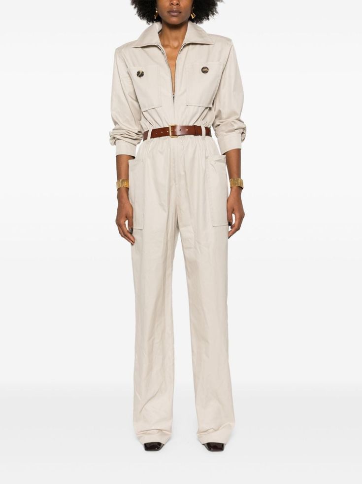 a woman wearing a white jumpsuit, standing in a studio and looking at the camera. she has her hair pulled back, and the jumpsuit is belted at the waist.