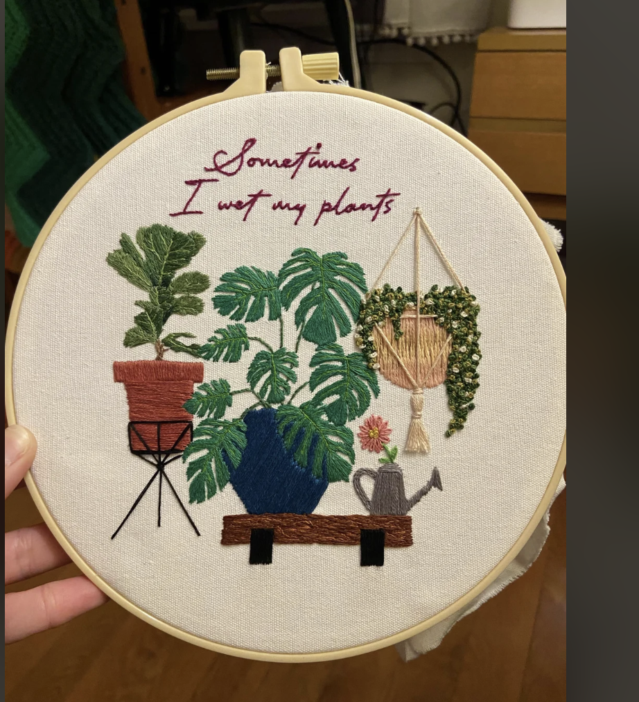 an embroidery hoop art piece featuring a collection of potted plants with the text "sometimes i wet my plants." embroidered above it. the plants are illustrated in various shades of green thread and include a monstera, a fiddle leaf fig, and a string of pearls. a watering can sits on a small bench below the plants. the background fabric is a light beige color and the embroidery hoop is made of natural wood. the photo is taken from an eyelevel perspective, capturing the entire embroidery hoop.