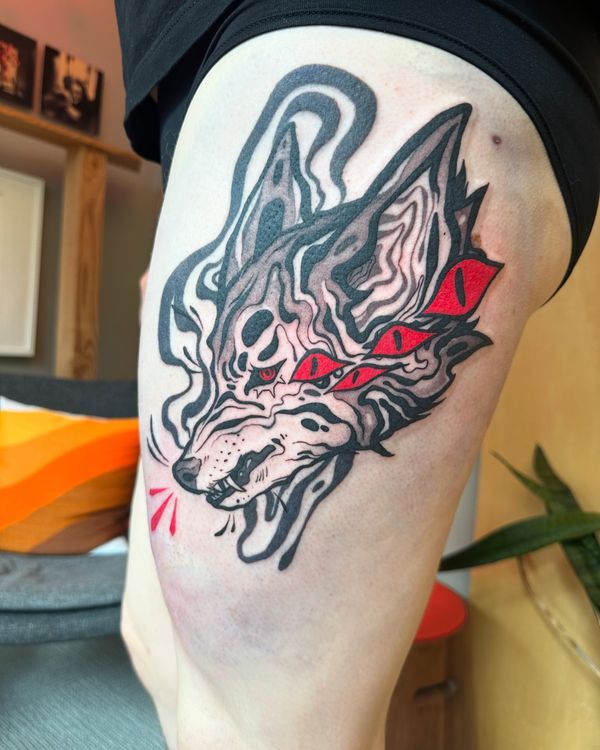 a closeup shot shows a person's thigh featuring a wolf tattoo with three red eyes. the tattoo is primarily done in black and grey ink, with the eyes providing a contrasting pop of red. the person is wearing black underwear. the background is slightly blurred.