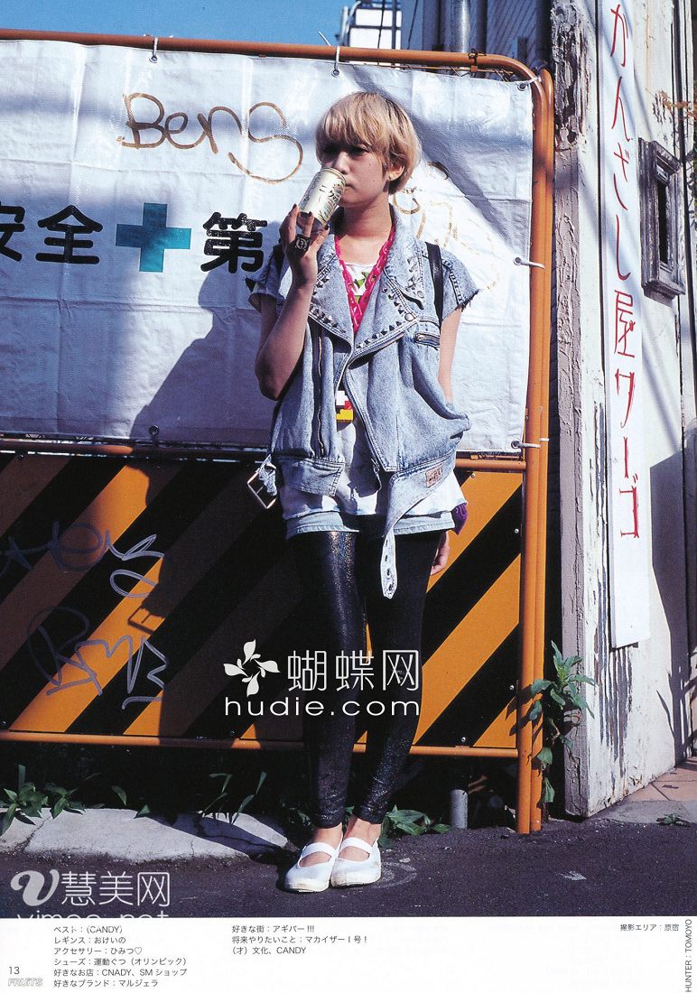 young woman with short blonde hair is standing in front of a construction barricade, posing for a street style photo. she's wearing a studded denim vest over a graphic tee, metallic leggings, and white shoes. she's holding a drink in her right hand and looking slightly offcamera. the background features the construction barricade with japanese text, highlighting the urban setting. the image is brightly lit, likely with natural daylight, creating a casual and candid feel.