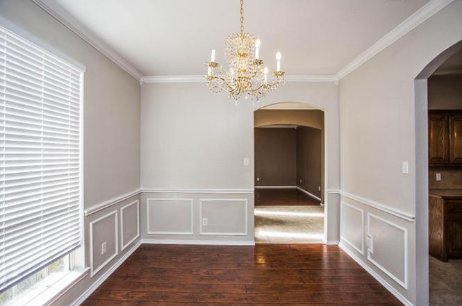 interior shot of a room with dark wood floors, light gray walls, white molding and wainscoting, and a gold chandelier hanging from the ceiling. there is a window with blinds on the left, and an arched doorway leading to another room in the background. to the right is an opening to a kitchen with dark wood cabinets.