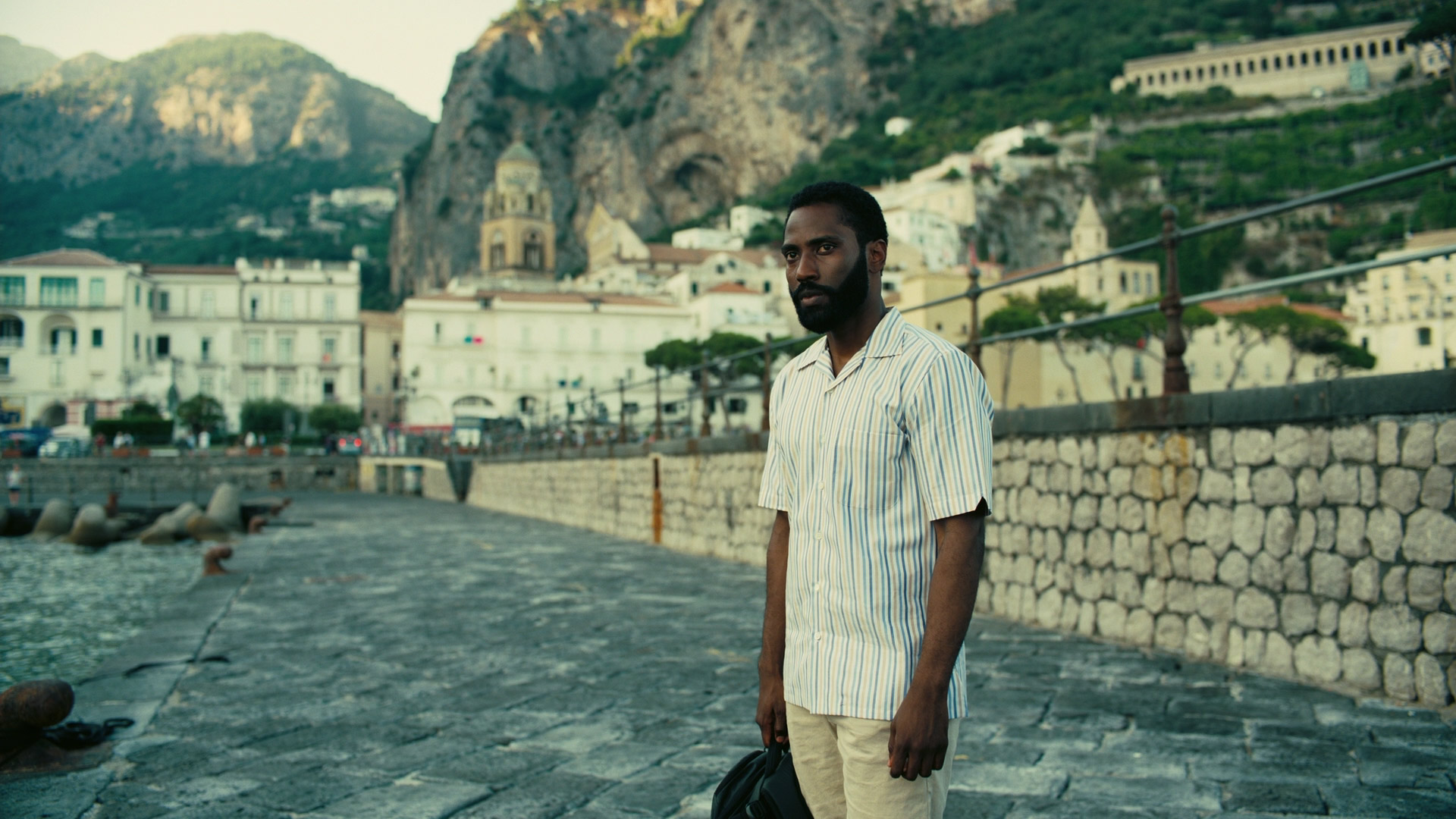 a man stands on a stone pathway along a waterfront in a town. the man is wearing a striped shirt and tan pants, and is looking at the camera with a thoughtful expression. he is holding a black bag. there are buildings and mountains in the background.