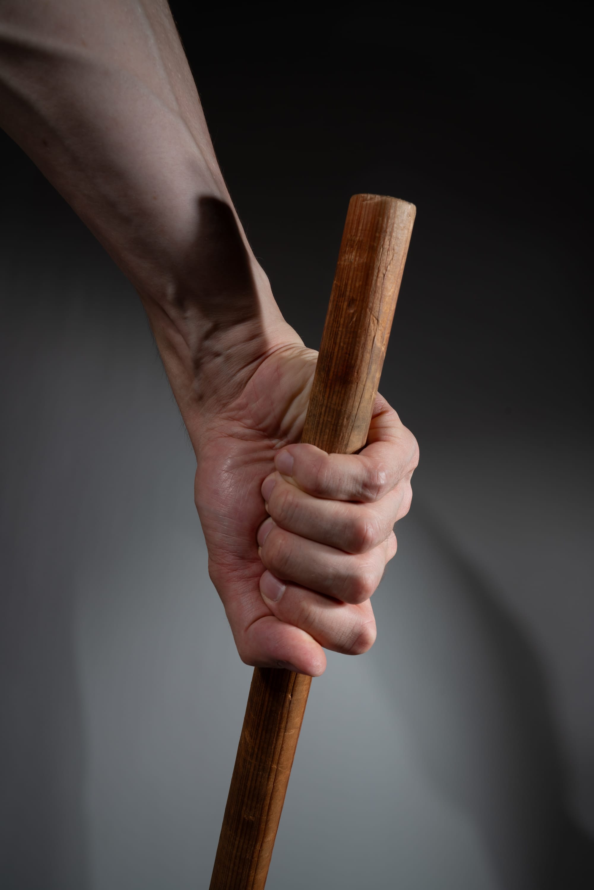 a man's hand tightly gripping a wooden stick against a gray background. the image emphasizes the strength and tension in the hand and forearm, highlighting the texture of the skin and wood.