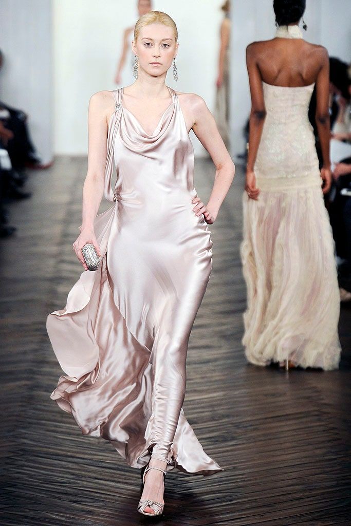 a model walks the runway in a silky champagnecolored gown with a cowl neck, holding a small silver clutch. she wears drop earrings and matching strappy heels. another model in a white gown walks behind her. they are on a dark wooden runway with an audience on either side.
