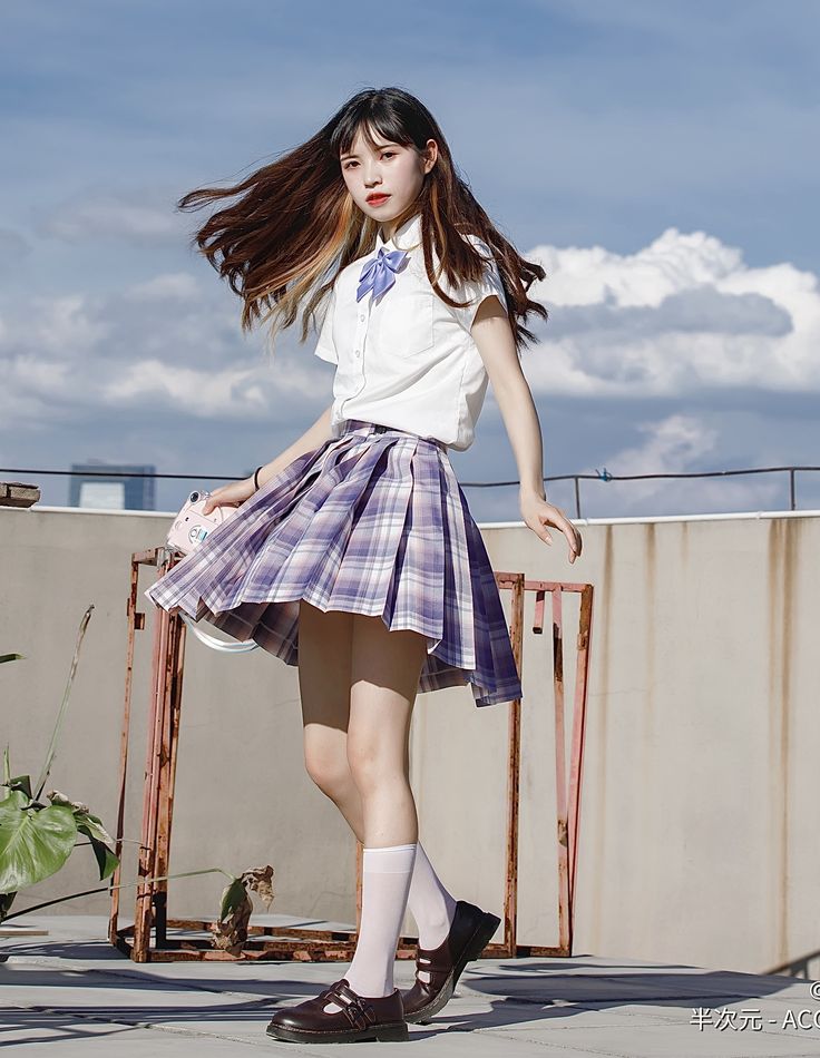 the image shows a young asian girl, possibly a high school student, dressed in a japanesestyle school uniform.  she's wearing a white shortsleeved shirt with a light purple bow tie, a purple and white plaid pleated skirt, white kneehigh socks, and brown mary jane shoes. her long brown hair is slightly windblown. she's posing on a rooftop against a backdrop of a partly cloudy sky. the overall style is cute and youthful, suggesting a fashion or editorial photoshoot.