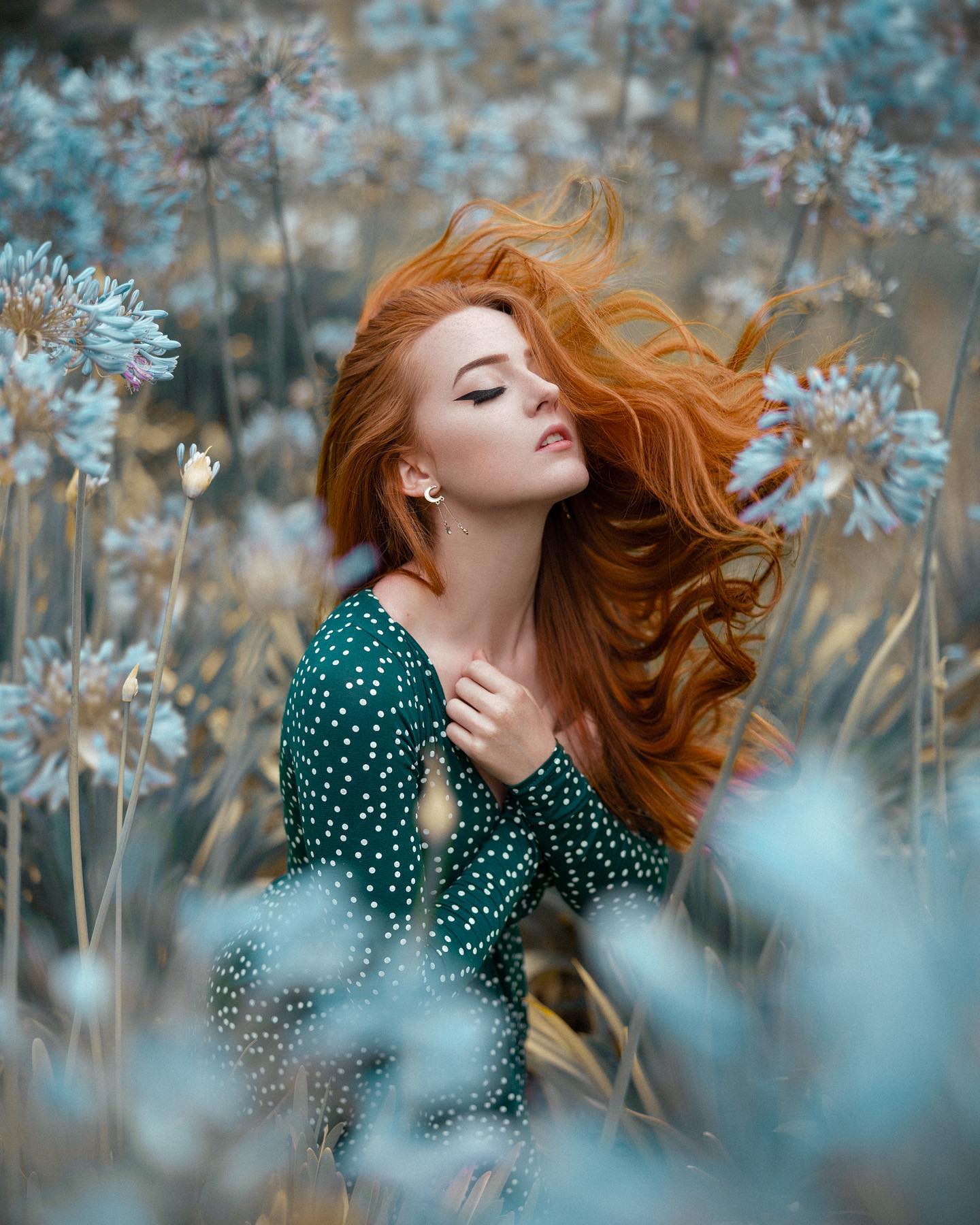 a woman with long, flowing red hair and a green polka dot dress is surrounded by tall, bluetoned flowers. she is looking up slightly with her eyes closed, her hands gently clasped at her chest. the photo is taken at eye level, with soft, natural lighting, creating a serene and peaceful atmosphere.