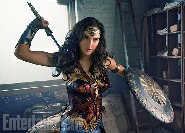wonder woman, played by gal gadot, is shown in costume holding a sword and shield in a warehouse setting. she is facing towards the right of the frame and ready to fight. she has a determined look on her face.