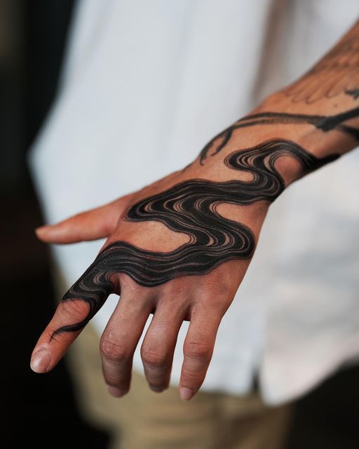 a closeup shot shows a man's hand and forearm adorned with an intricate black tattoo. the tattoo features a pattern of wavy lines, creating an abstract design that covers the back of the hand and extends onto the forearm. the man's skin tone is visible beneath the tattoo, and his fingers are slightly extended. the background includes a white shirt and a glimpse of beige pants.