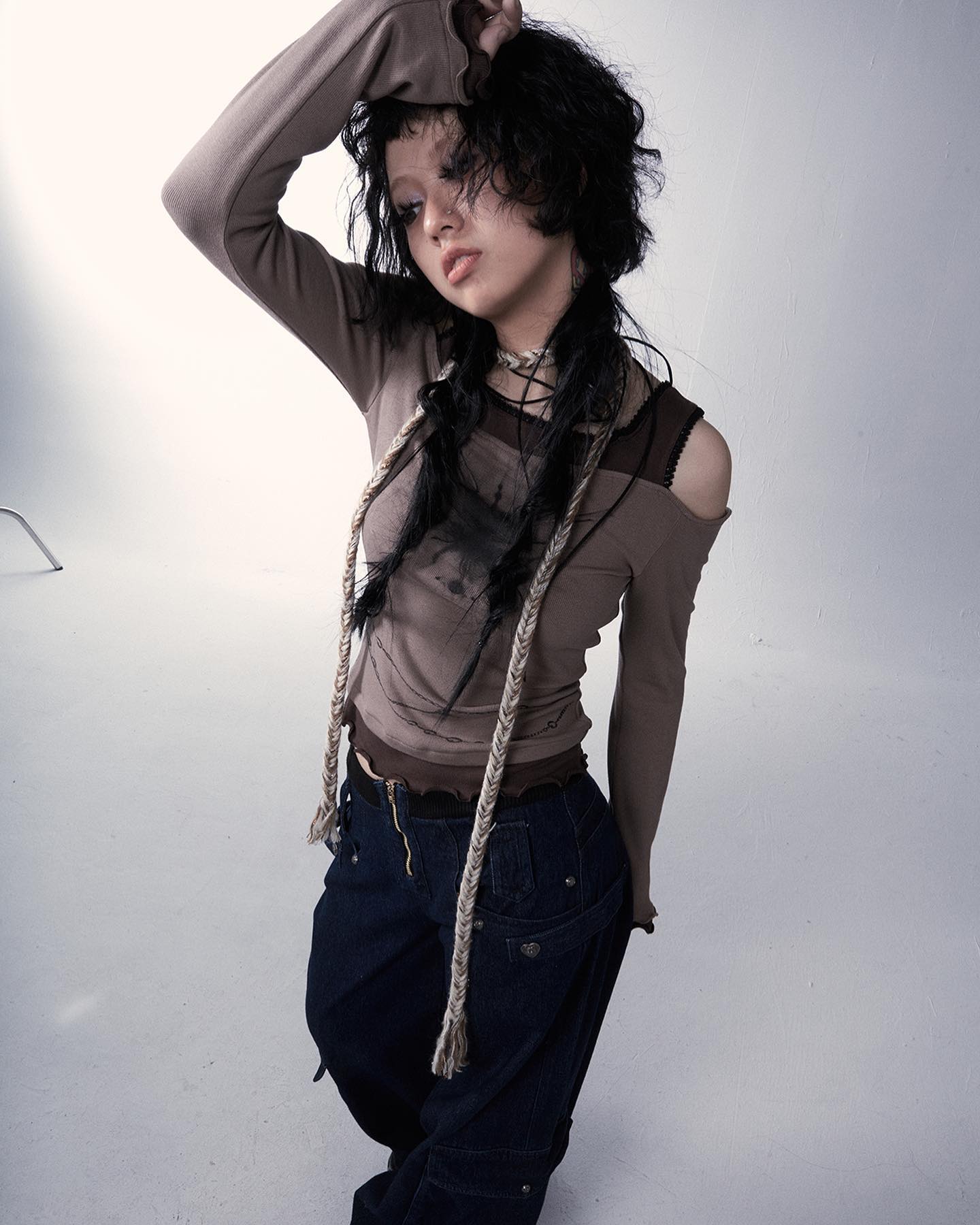 the image shows a young woman, possibly a fashion model, posing against a plain grey backdrop. she is wearing a longsleeved, taupecolored top with an offtheshoulder design.  the top has a distressed or slightly worn look. her bottom is a darkwash pair of baggy jeans with visible pockets and detailing.  she's also accessorized with a light beige rope necklace/accessory that drapes down her chest, and a dark choker.  her dark hair is styled in loose, slightly tousled waves. she has a somewhat serious and intense expression. the overall style is edgy, grunge, and alternative fashion.  the lighting and photography style enhance the moody and fashionable feel.