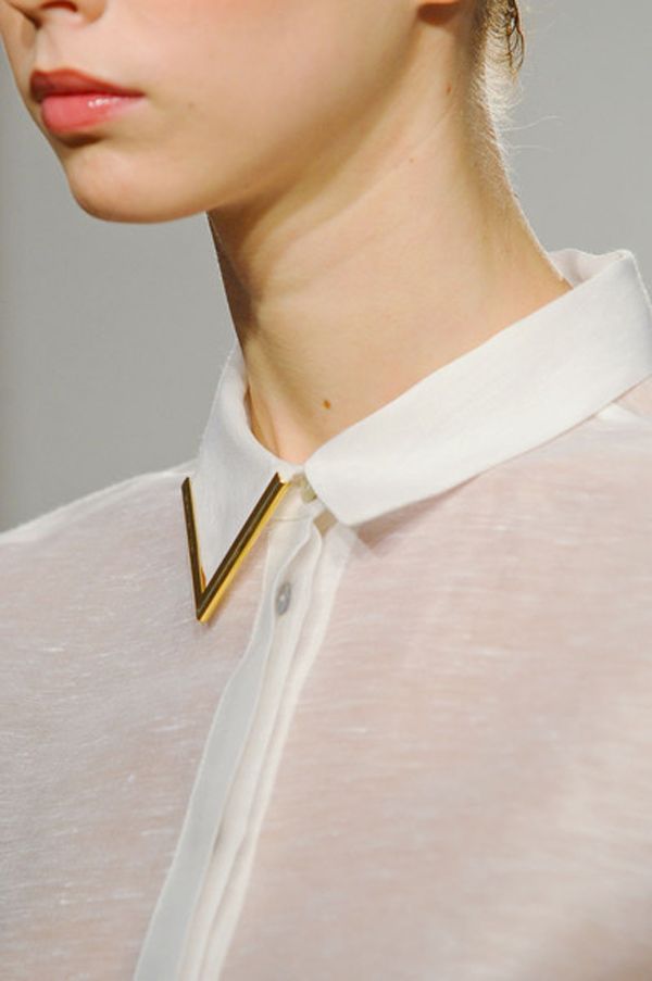 closeup of a woman model wearing a sheer white shirt with a gold, vshaped collar accessory. the image focuses on the detail of the shirt and jewelry, highlighting the minimalist and elegant style. the woman's face is partially visible, showcasing her soft makeup and subtle expression. the background is blurred and out of focus, drawing all attention to the model and her attire.