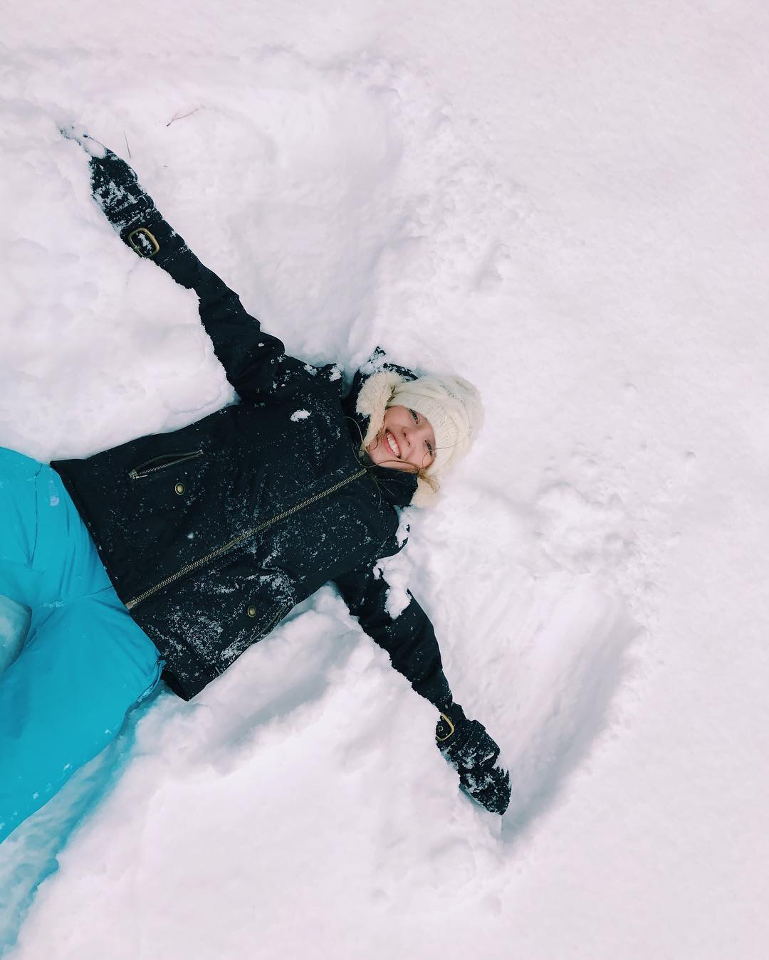 a young woman lies in the snow, making a snow angel. she is smiling and appears to be enjoying herself. she is wearing a black jacket, blue pants, and a white hat and gloves. the snow is fresh and fluffy, and the snow angel is clearly visible. the image is bright and cheerful, capturing a moment of winter fun.