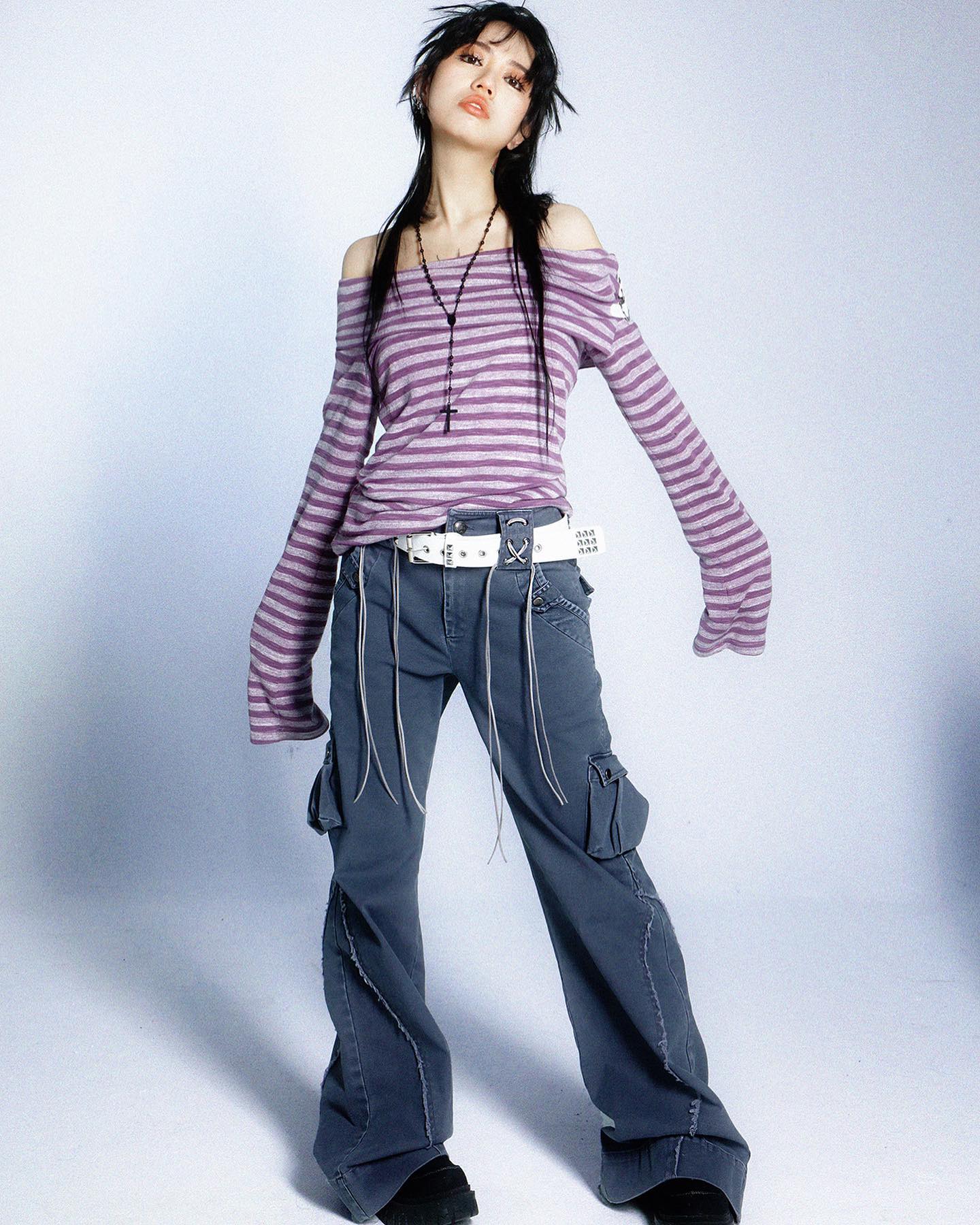 the image is a fulllength studio shot of a young asian woman modeling a casual outfit. she has long dark hair and is wearing a lilac and white striped, offtheshoulder longsleeved shirt, paired with loosefitting, gray wideleg cargo pants and a silver belt. a simple black cross necklace is visible around her neck. she is posing slightly turned to her side, showing off the outfit’s details. the background is a plain light blue or gray, providing a clean contrast to her clothing. the overall style is reminiscent of y2k or alternative fashion.