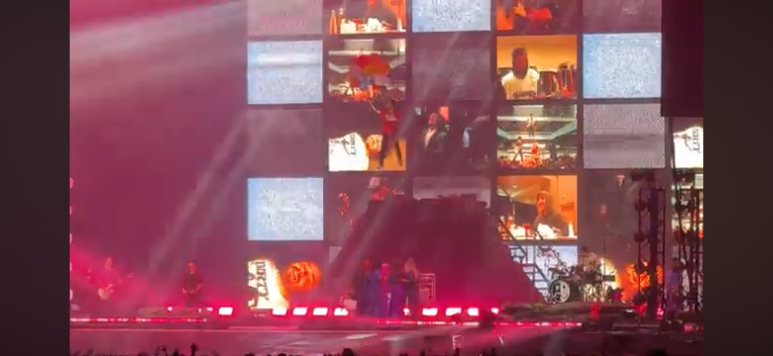 a live concert performance on a stage with a large screen displaying a video of a band. the stage is lit up with colorful lights and the band is playing their instruments. the audience is gathered in front of the stage, cheering and clapping along. the energy of the performance is palpable.