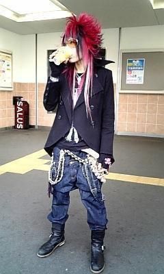 a person with long red and black hair stands on a gray surface. they are drinking from a container with their right hand. the person is wearing black sunglasses, a black jacket, blue jeans and black boots. a long chain hangs from their pants. they are inside a building that has white walls and tile flooring. a black storage container sits in the background.