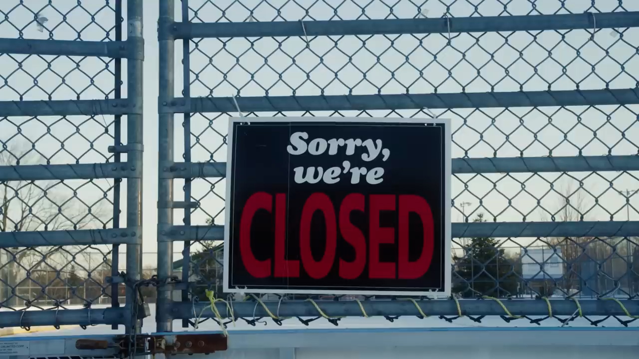 a chain link fence with a closed sign on it. the sign is black with white and red letters. the ground is covered in snow.
