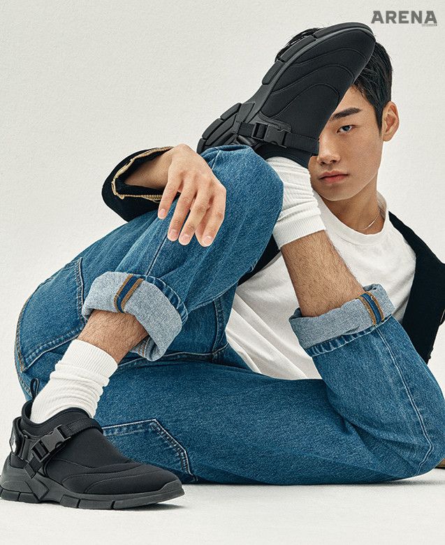 a man wearing blue jeans, a white shirt, and black sneakers is sitting with his legs crossed and looking at the camera.  he has his foot resting on his knee and he has a slight smile on his face.  the background is white.