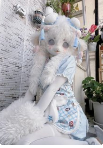 the image shows a person wearing a fullbody, fluffy white costume resembling a lamb or sheep character. the costume features large ears, pastel blue and white clothing, and exaggerated features. the person is sitting down, their face obscured by the costume's head. the setting appears to be indoors, possibly on a balcony, with tiled walls and potted plants visible in the background. the lighting suggests natural daylight.