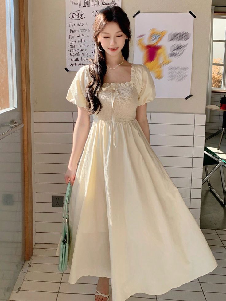 a young woman is posing indoors, wearing a pale yellow puff sleeve midi dress with a square neckline and a gathered bodice. she is also carrying a green shoulder bag and wearing white sandals. the background features a door and a wall decorated with a cafe menu.