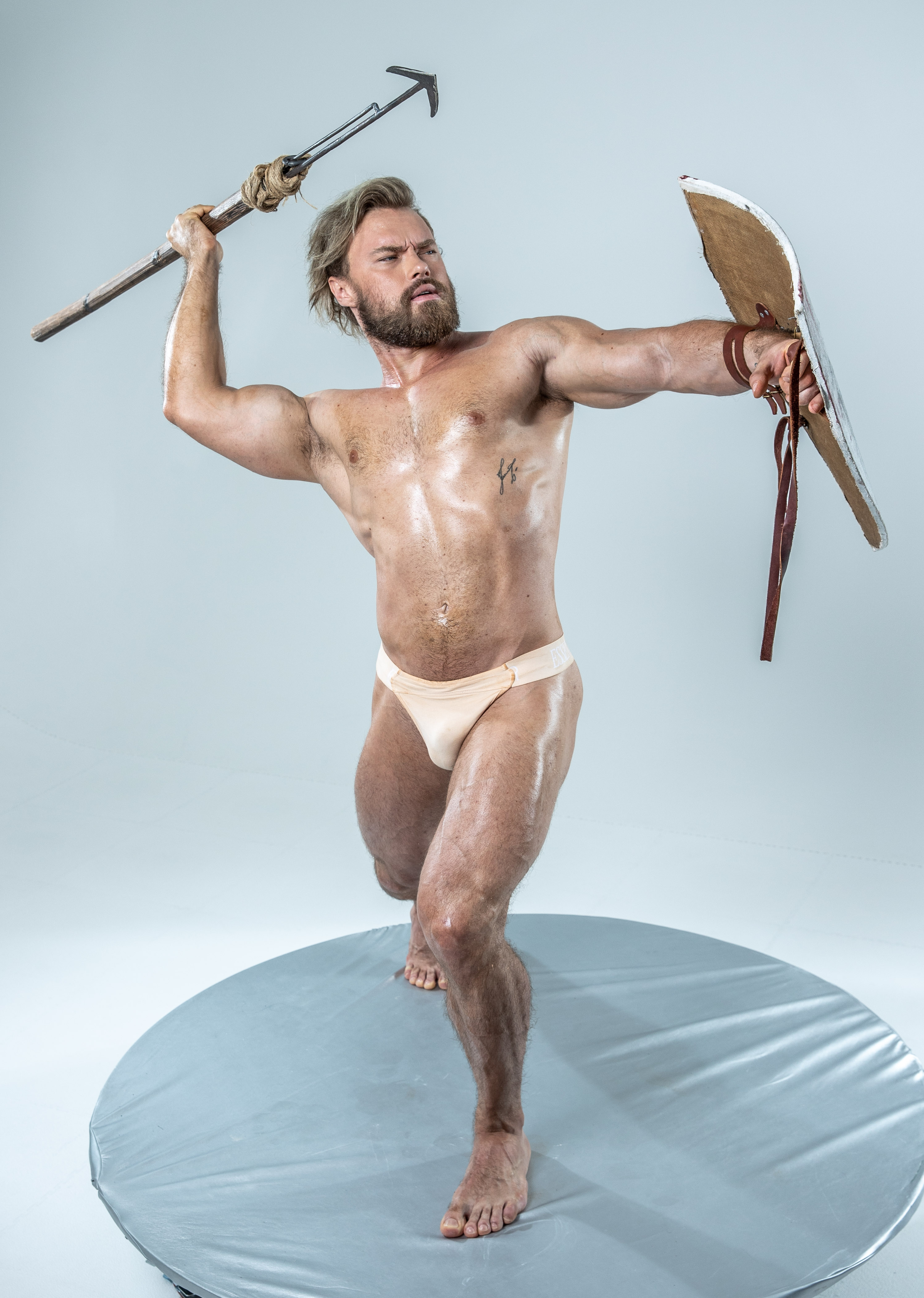 a muscular man, embodying the spirit of a spartan warrior, stands poised for battle within a studio setting. his weapon, a rudimentary spear, is grasped firmly in his right hand, raised midswing, while a weathered shield adorns his left arm, prepared to deflect any incoming attack.  his stance speaks of power and readiness, muscles tensed, gaze fixed on an unseen adversary. the bright studio lighting accentuates the definition of his physique, highlighting the sweat glistening on his skin.