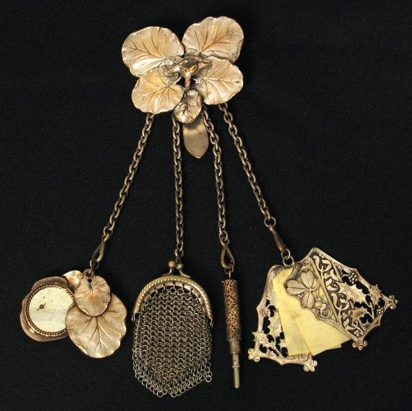 a chatelaine with a flower brooch at the top from which hang chains with a coin purse, pencil, notepad, and watch.