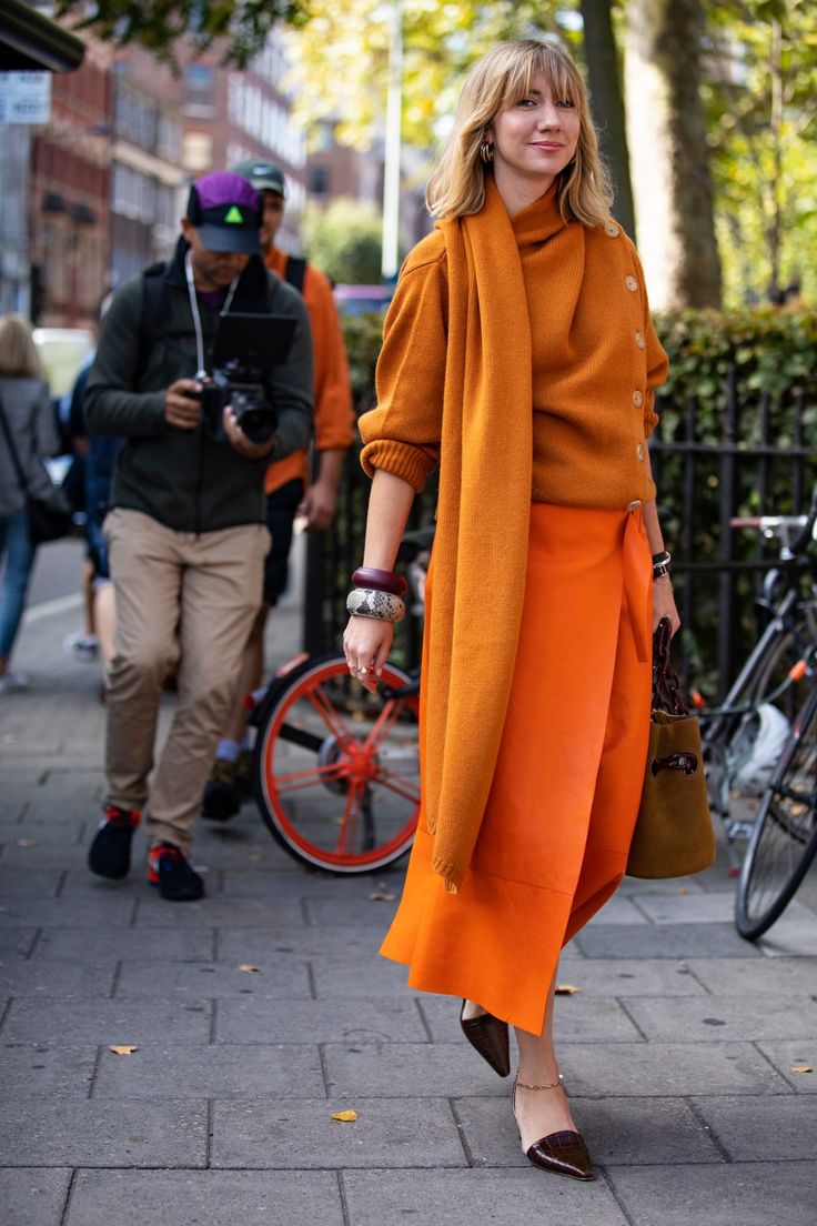 a woman in an allorange outfit walks down a city street. she is wearing an orange sweater, an orange scarf, an orange skirt and brown heels. she has a  brown purse over her shoulder and is wearing silver bracelets on one wrist. a photographer is capturing her style from the background.